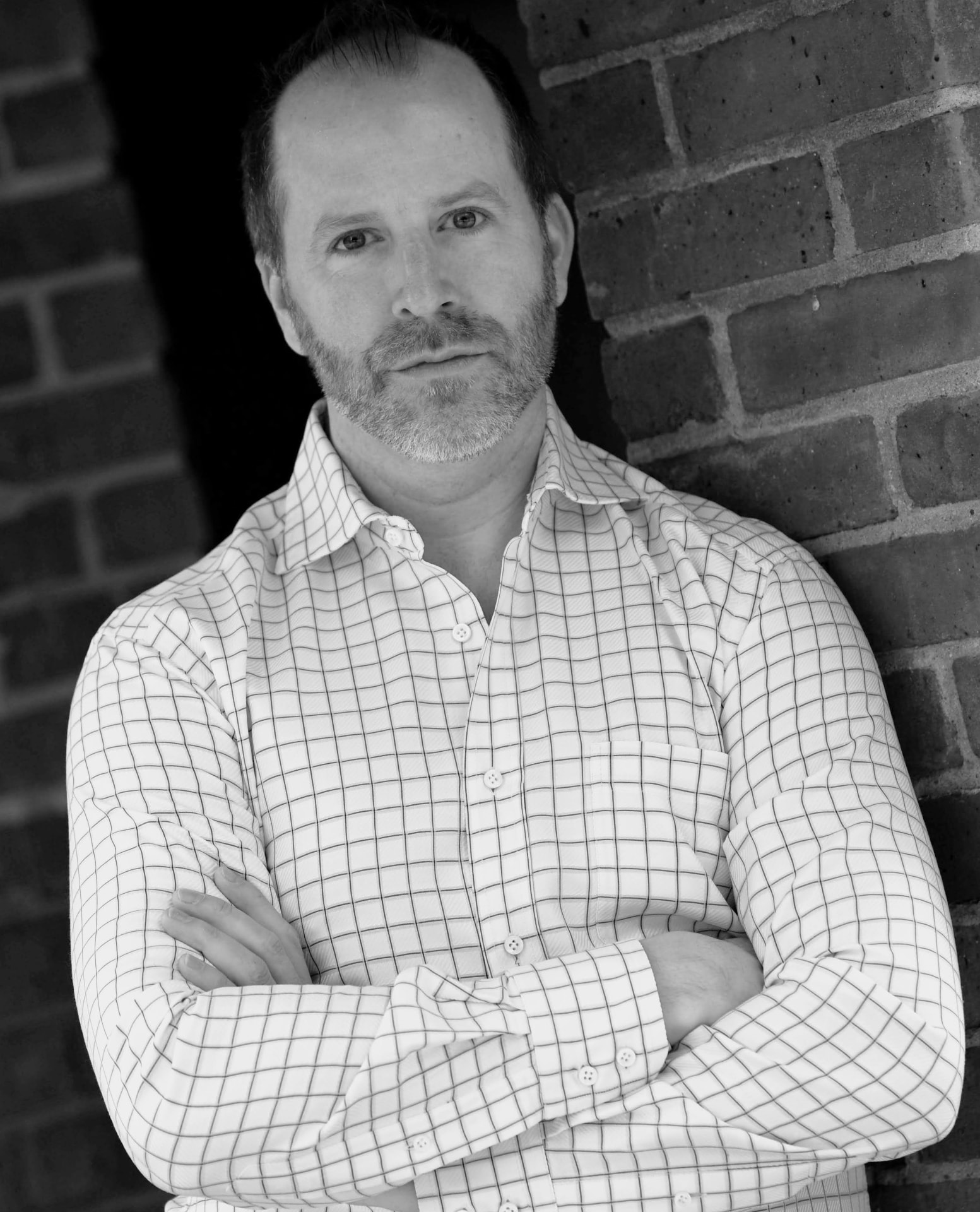 Black-and-white portrait of Joshua Whitaker, arms crossed, leaning on a brick wall